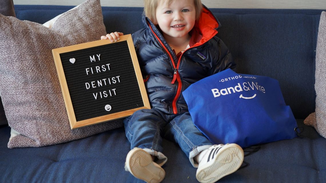 A toddler sitting in a big chair at a dental office, holding a sign that says, "My first dentist visit." He is smiling big and holds a Band & Wire merchandise bag.